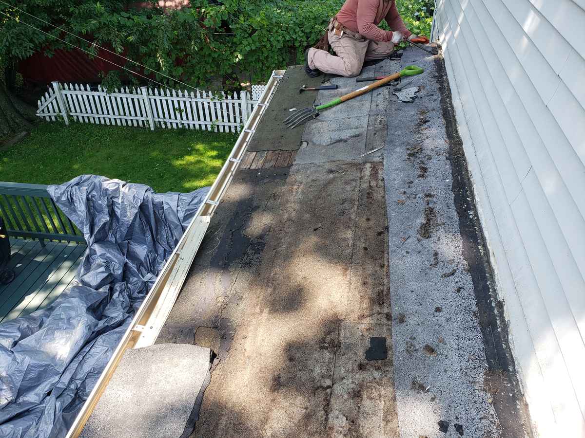 Roofer preparing the edge of a low-slope roof beside white siding, with tarps and tools
